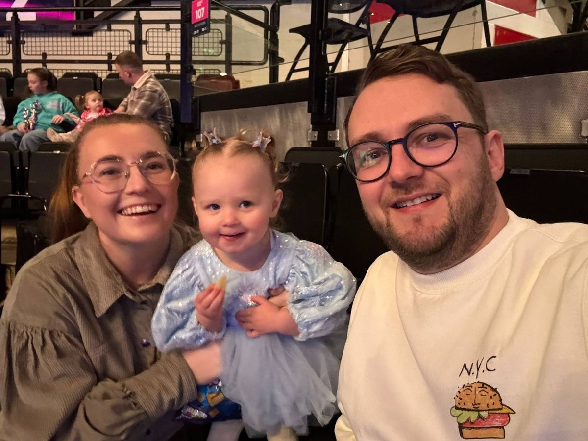 An image depicting a family of three, where two adults and a child are posing together in a cheerful manner, likely at a sporting event or a similar venue. The adults are both wearing glasses, and the child is wearing a blue long-sleeved outfit. The background shows a partially blurred view of an audience and a structure.