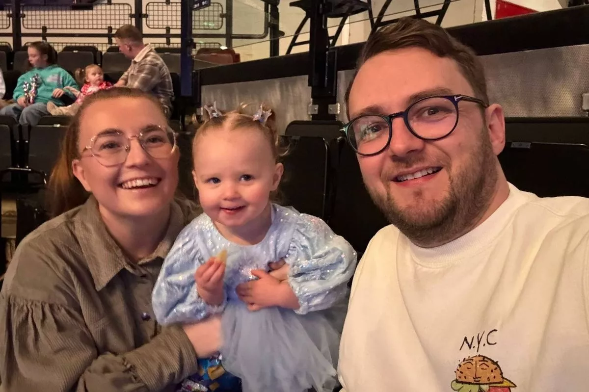A family of three, consisting of two adults and a child, are seated together in a stadium. The adults are wearing glasses and are smiling, while the child is holding a small object and also smiling. The background shows a blurred view of other spectators in the stadium.