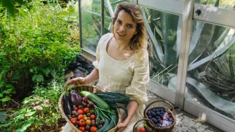 Ramona Jones A young woman with brown hair is wearing a flowy cream dress. She is smiling and is sitting by the edge of a greenhouse with a large wicker basket filled with fresh vegetables on her lap. In the background is a lush green garden. 