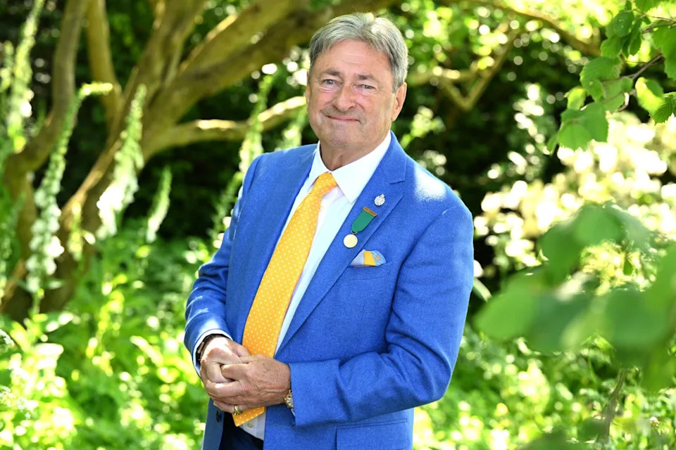 a picture of alan titchmarsh a man wearing a blue blazer, white shirt, and yellow tie, standing in front of a tree and flowers