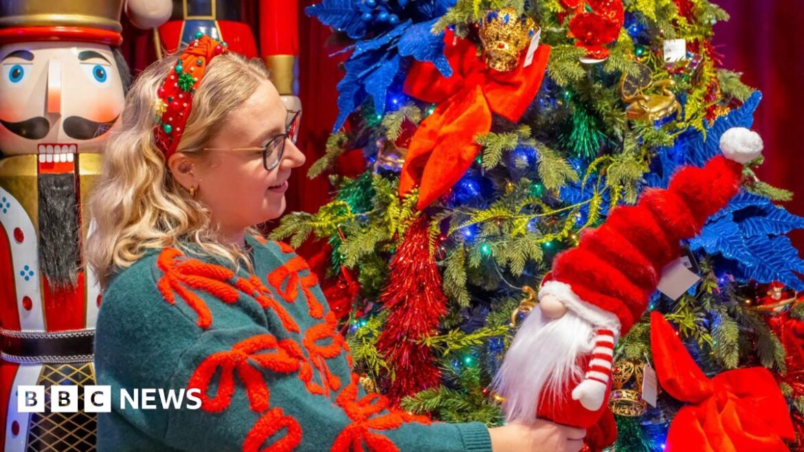 A woman with blonde hair wearing dark framed glasses and a green and red Christmas jumper is stood in front of a Christmas tree holding a toy father Christmas.