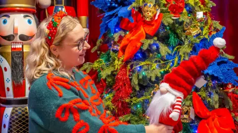 Blue Diamond A woman with blonde hair wearing dark framed glasses and a green and red Christmas jumper is stood in front of a Christmas tree holding a toy father Christmas.
