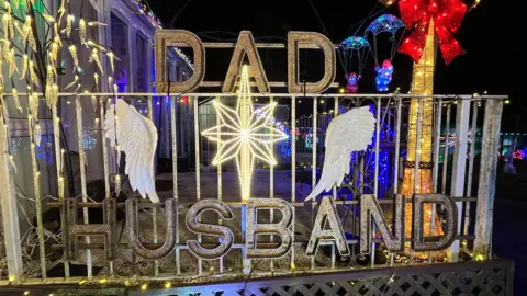 A metal sign that reads DAD HUSBAND with two angel wings, a star in the centre and strings of lights.