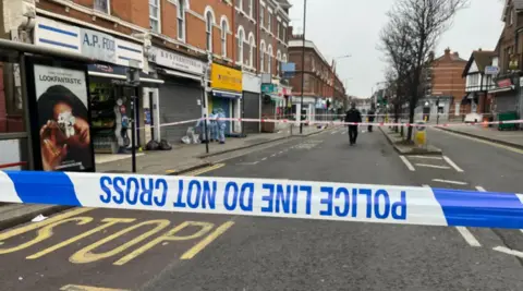 Police cordon tape on a London road. A police officer is seen in the distance.