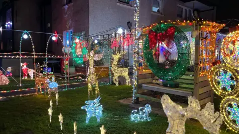 An array of Christmas decorations and lights in a garden. There are reindeer, holly wreaths and coloured garlands.