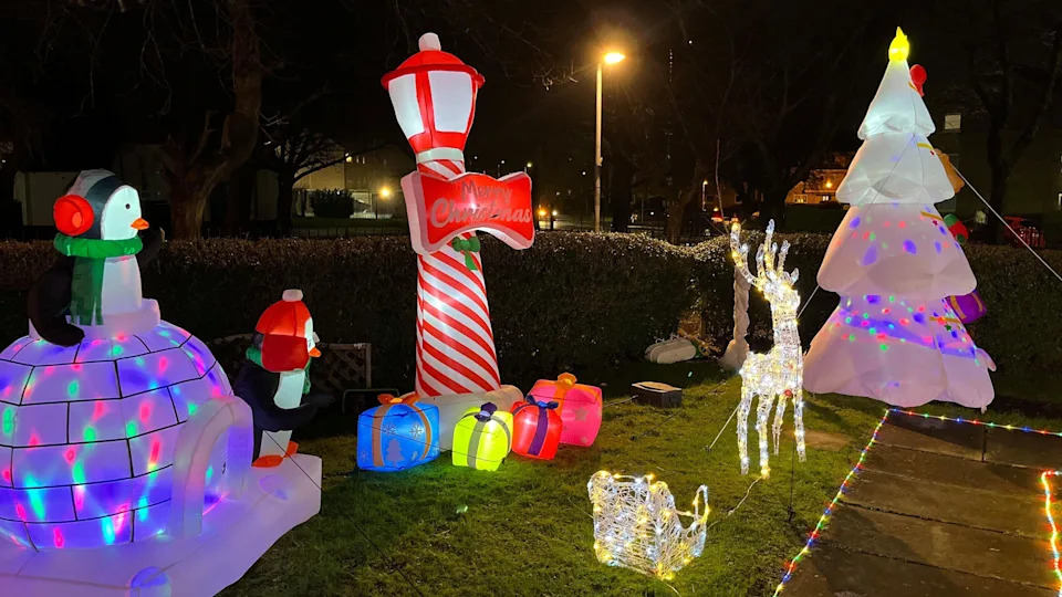 Inflatable Christmas decorations on the lawn. There is a multi-coloured igloo with penguins, a red and white striped lamppost with parcels around its base and a white Christmas tree. A reindeer lights also sits on the grass.