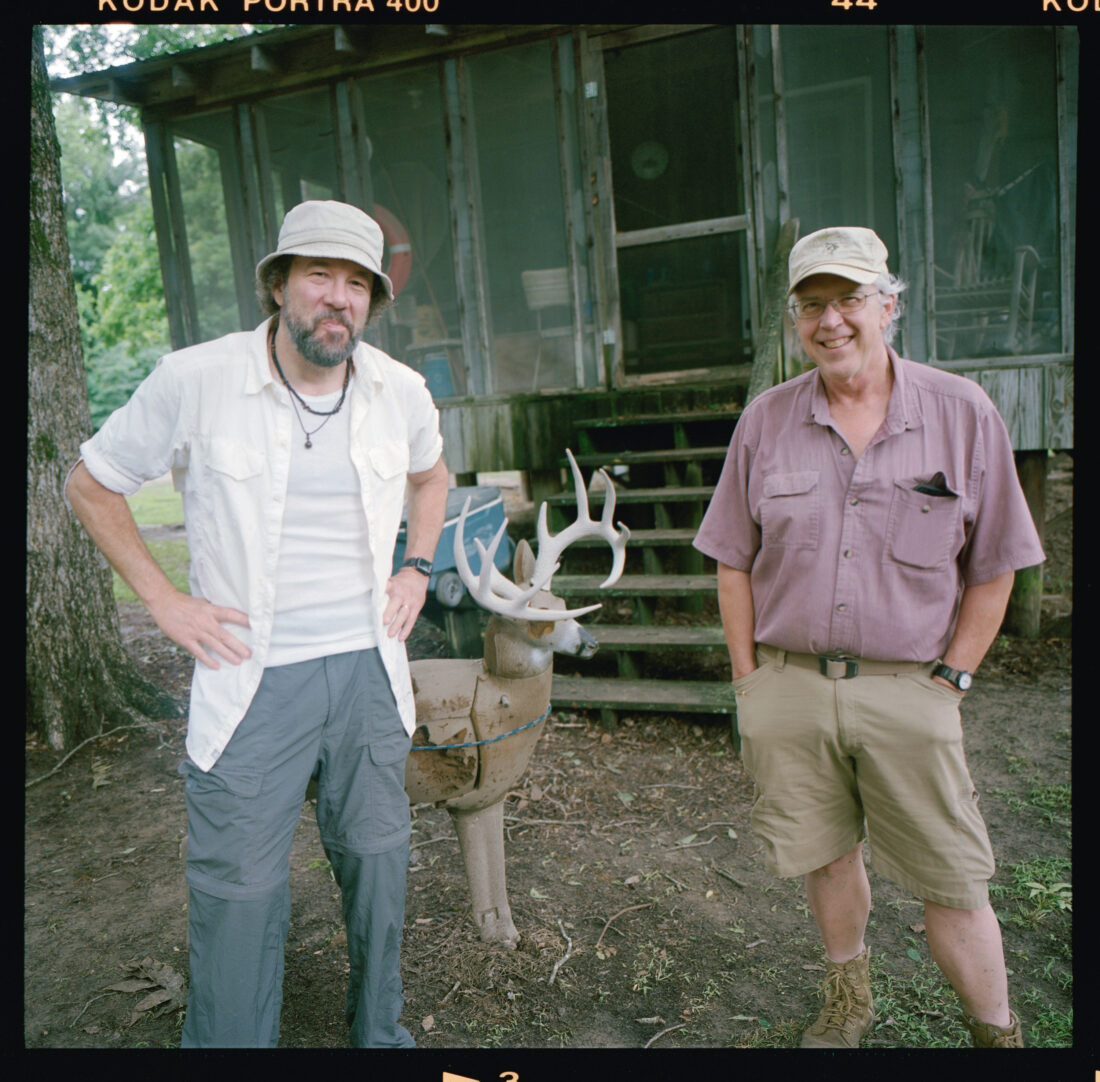 Two men stand in front of a hunting cabin