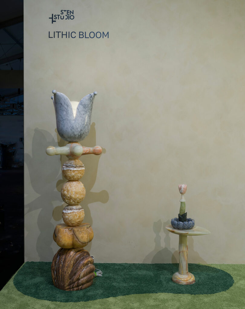 Two abstract stone sculptures by Sten Studio stand on green carpeting against a beige wall, with the text "STEN STUDIO LITHIC BLOOM" displayed above them.