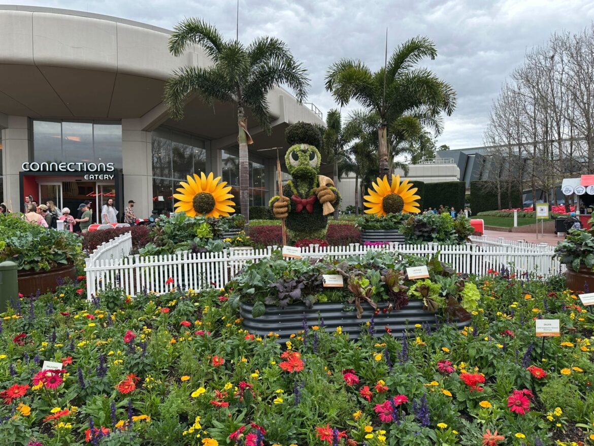 A charming topiary of a character gently cradling a butterfly stands amidst a vibrant garden of sunflowers and colorful blooms, all framed by a white picket fence. In the background, "Connections Eatery" beckons with its inviting presence.