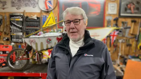 BBC A man in his 70s stands in front of a wheelbarrow, adorned in Christmas decorations including bows and baubles, in his garage. There are tools neatly hung all over the wall. He is wearing a white turtleneck, a black and grey coat, and black-rimmed glasses.