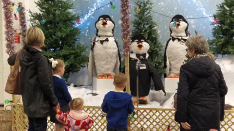 Gordale garden centre Three children with blonde hair and two older women ared standing in front of a display of three singing penguins.