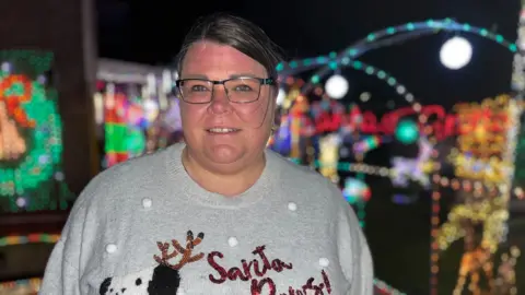 BBC Sandra Lee Myles wearing a grey Christmas jumper standing in front of the Christmas lights in her garden.