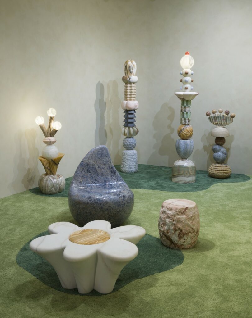 A room with green carpet features abstract sculptures and Sten Studio furniture made of stone, including a flower-shaped table, a blue chair, and several totem-like structures with spherical elements.