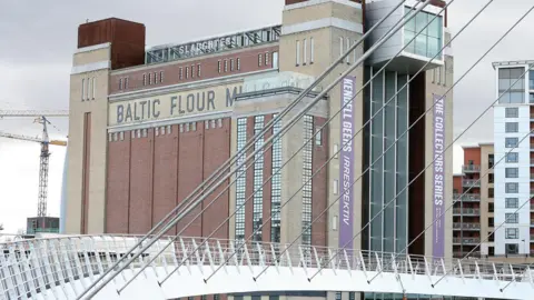 PA Media The Baltic Centre For Contemporary Art is a tall red-brick building on the River Tyne. Near the top, black bricks are laid in way that spells Baltic Flour Mills. The white Millennium Bridge is in the foreground of the photograph.
