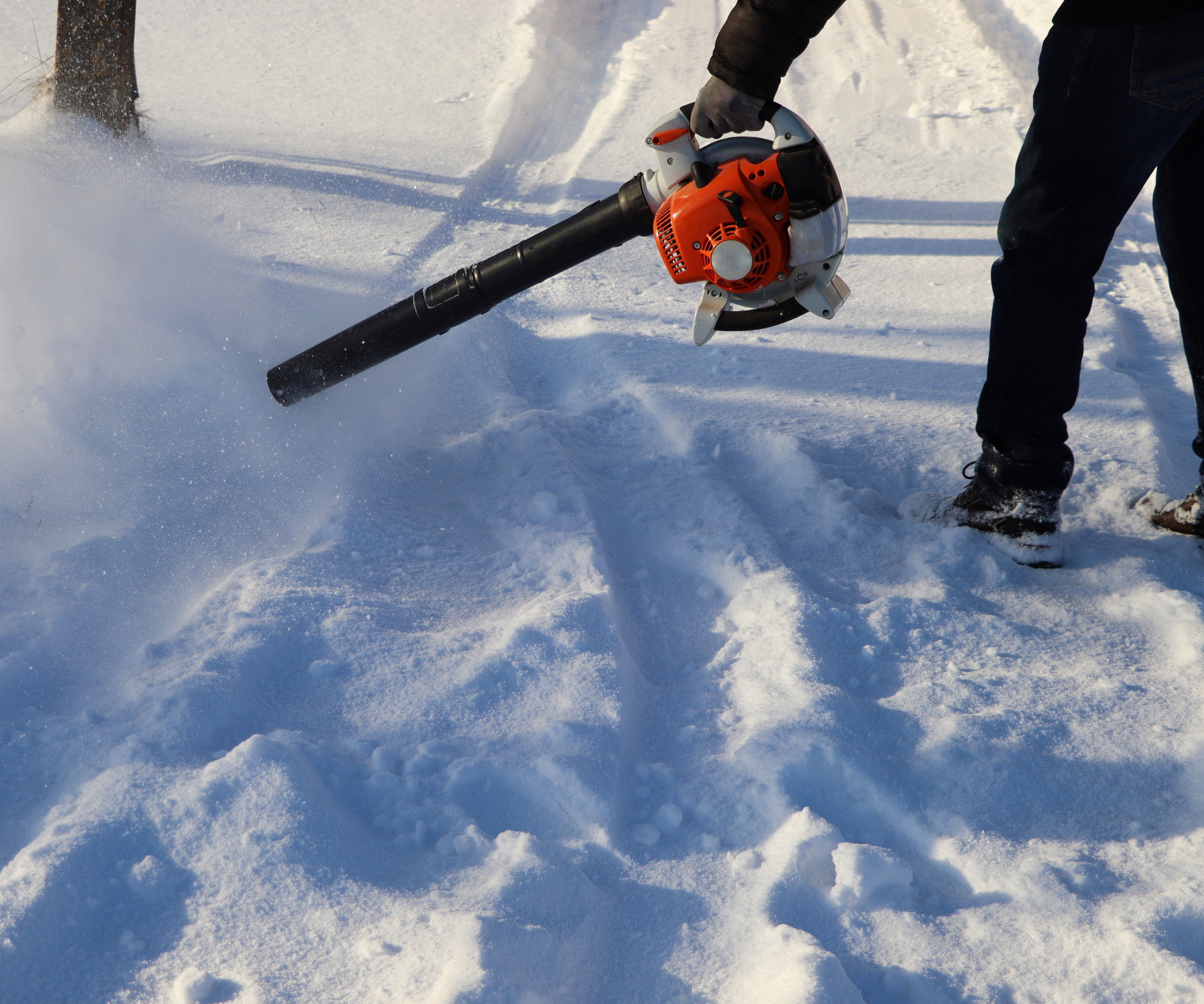 A person is using a handheld leaf blower to clear thick snow off a sidewalk
