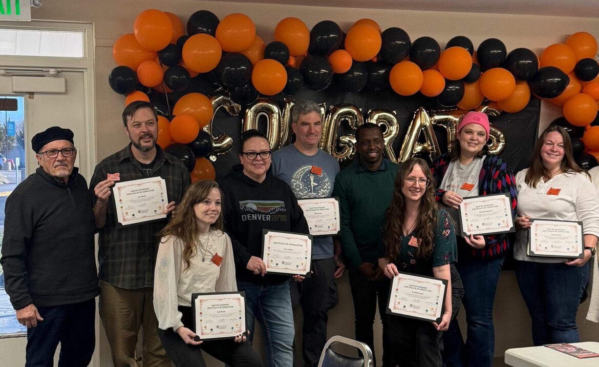 A group of people holding certificates stand in front of a wall decorated with orange and black balloons spelling “Congrats.”