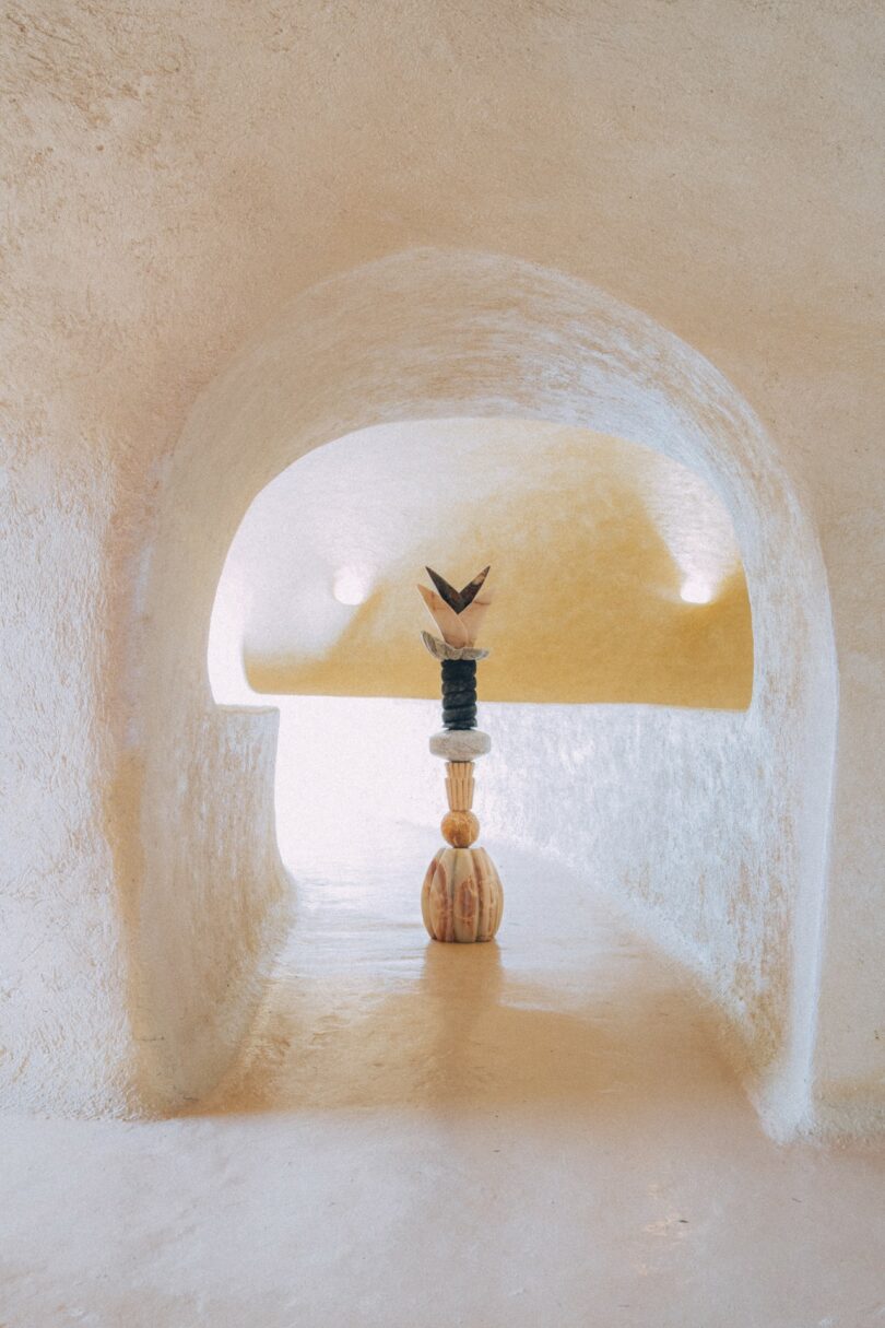 A sculptural art piece with stacked geometric shapes stands in the center of a smooth, beige, arched hallway with soft lighting