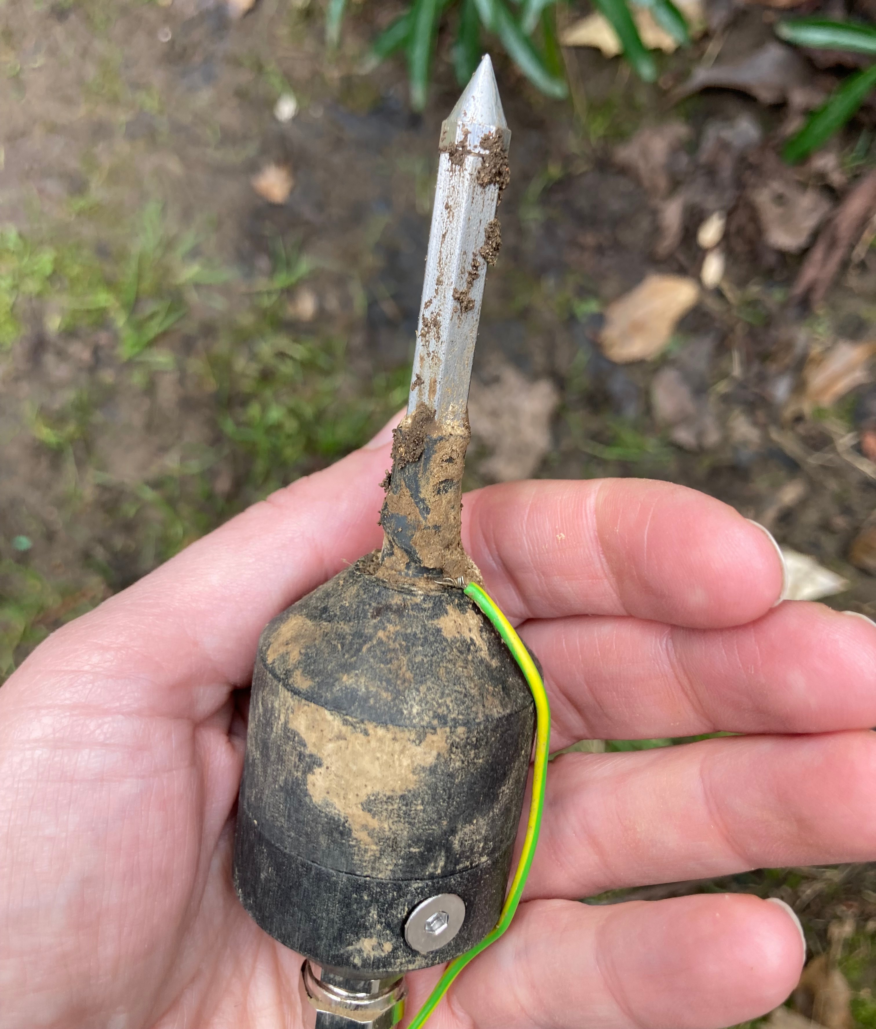 A hand holds a soil sensor device with a pointed metal tip covered in dirt.