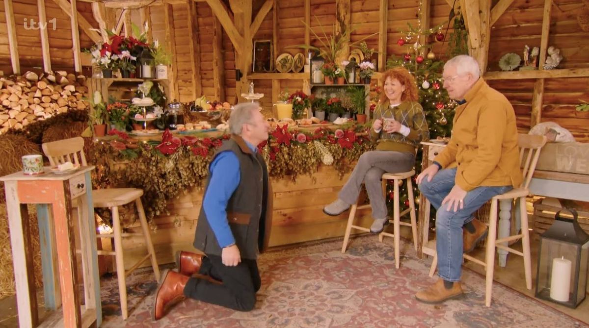 In a warmly decorated room, three individuals are engaged in a conversation. The setting features a wooden bar adorned with festive decorations, including garlands and seasonal ornaments. Two of the individuals are seated on stools, while one is kneeling on the floor. The ambiance suggests a cozy and festive gathering.