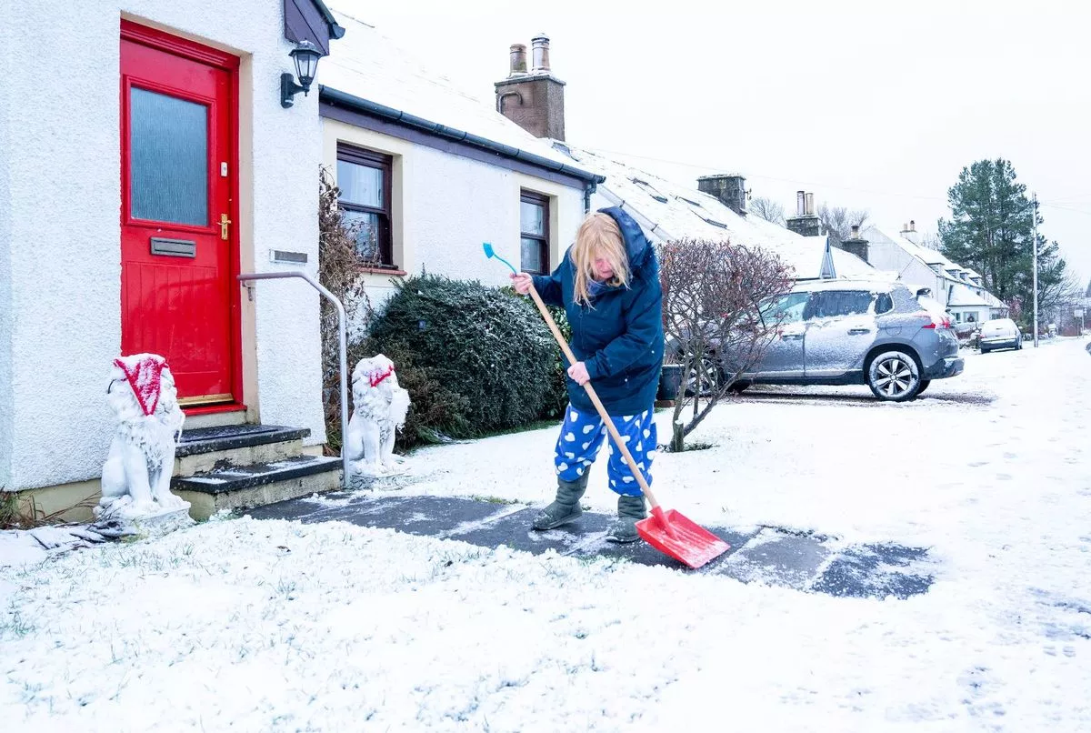 An individual is clearing snow from the front of a house using a shovel. The house features a red door, and there are snowmen and a parked car visible in the background.