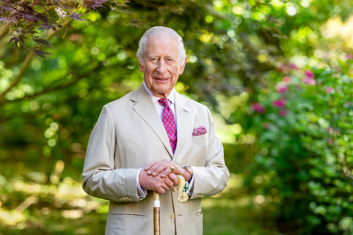 An older gentleman with white hair, wearing a light-colored suit and a pink tie, is standing outside and smiling while posing for a photograph. The background features lush greenery.