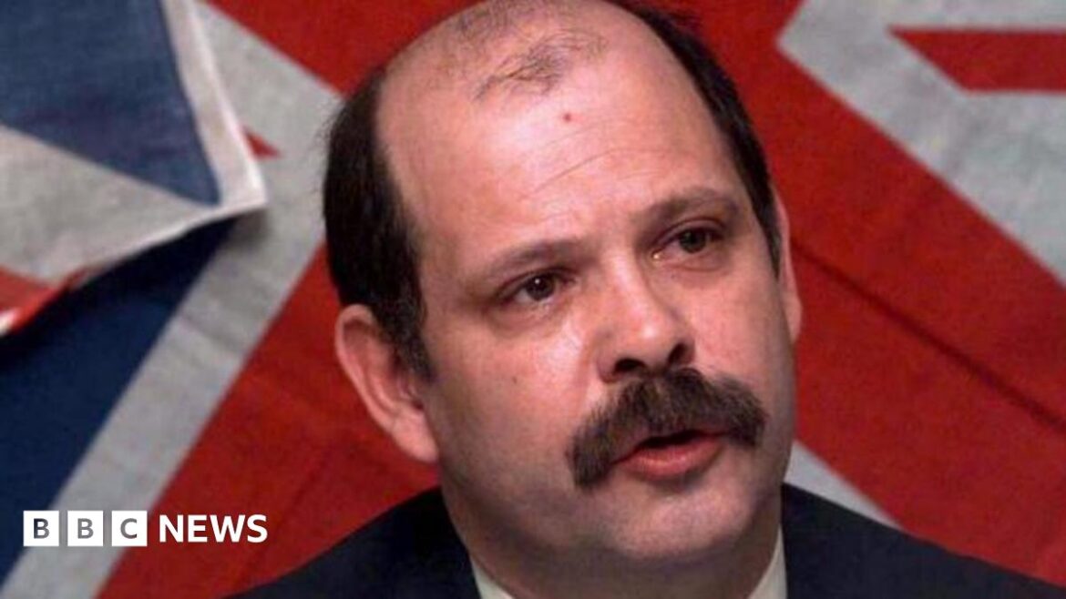 The late David Ervine, pictured from the shoulders up. He is balding and has dark brown hair and a brown moustache. there is a Union Jack flag on the wall behind him.