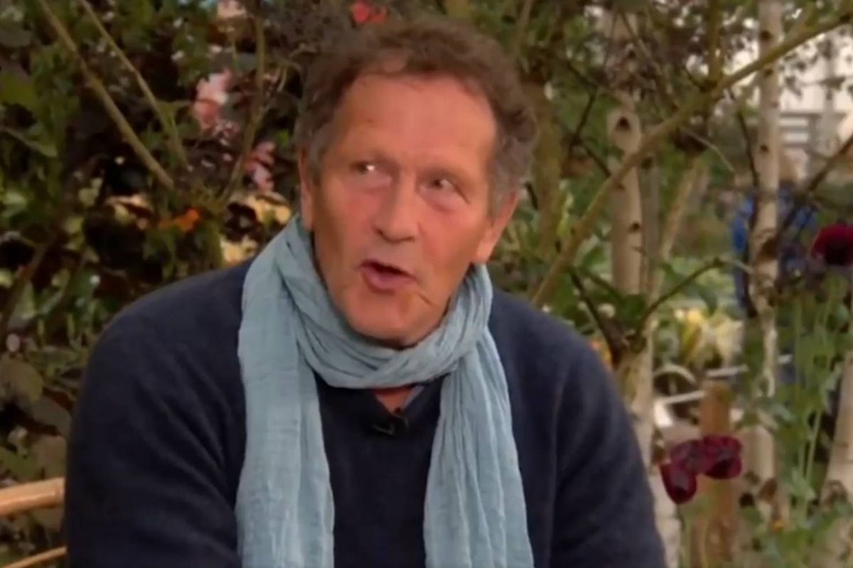 An individual, dressed in a dark-colored top and adorned with a light blue scarf, is seated in an outdoor setting. The backdrop features lush greenery and partial views of what appears to be a building. The person's expression suggests engagement in a conversation or interview.