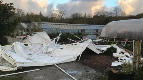 Lanjeth Nursery and Water Gardens A white marquee is upside down and all the poles are twisted and broken,  behind that is a poly tunnel which has remained unscathed and a 30 metre long glasshouse that has panes missing in it.  