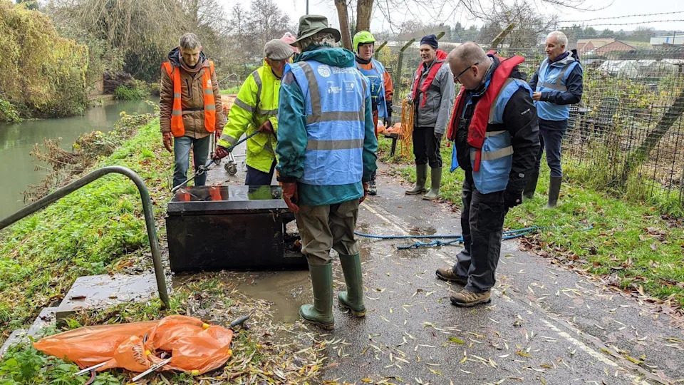 A group of litter pickers wearing blue, yellow and orange high vis vests,  standing on tarmac path on top of a grassy embankment by the side of a river. They appear to have retrieved an old electrical appliance which is lying on the path with a length of blue rope attached to it.