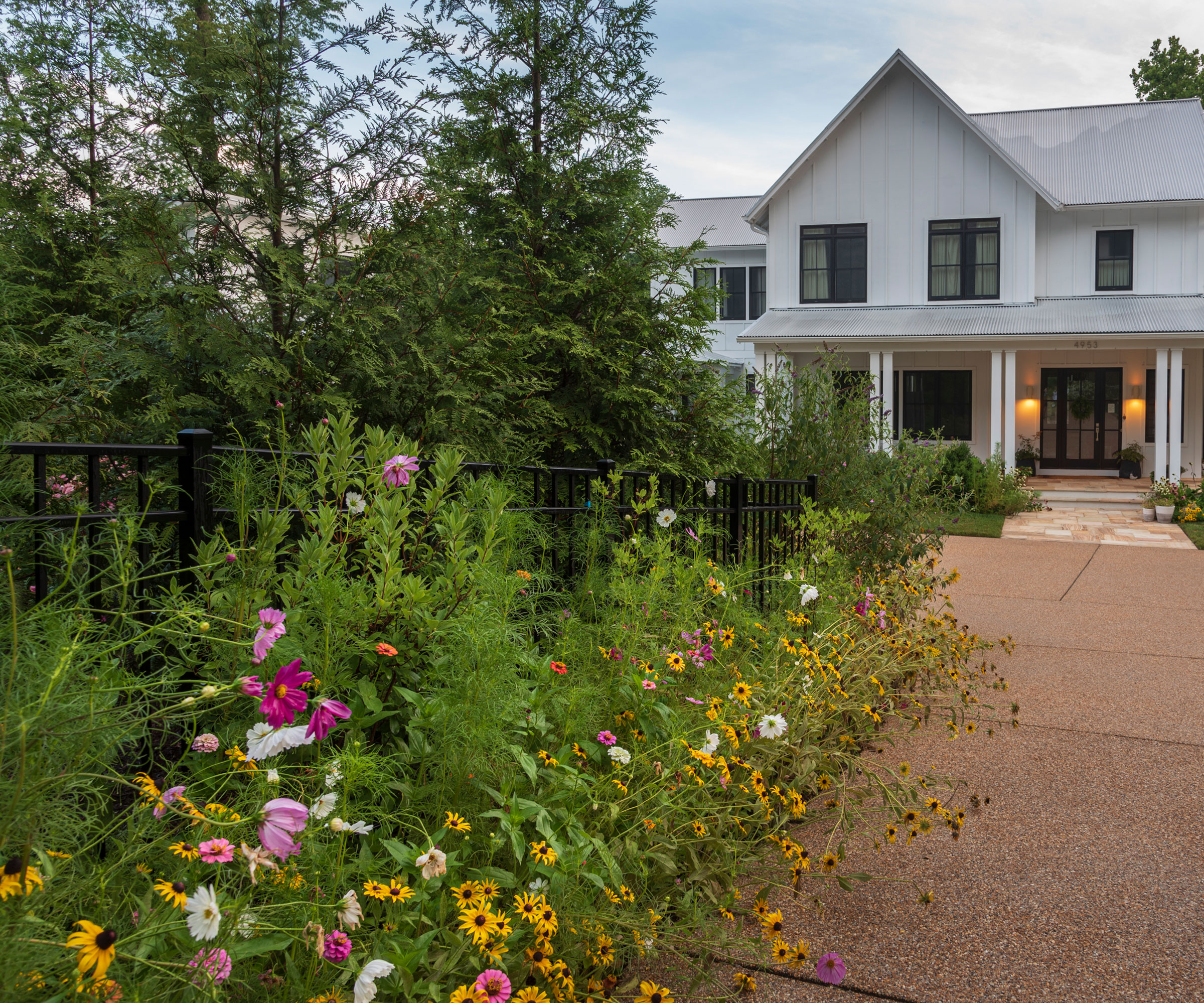 mosaic style planting in a long bed in the front garden of a house with a wide garden path and trees and house in the background