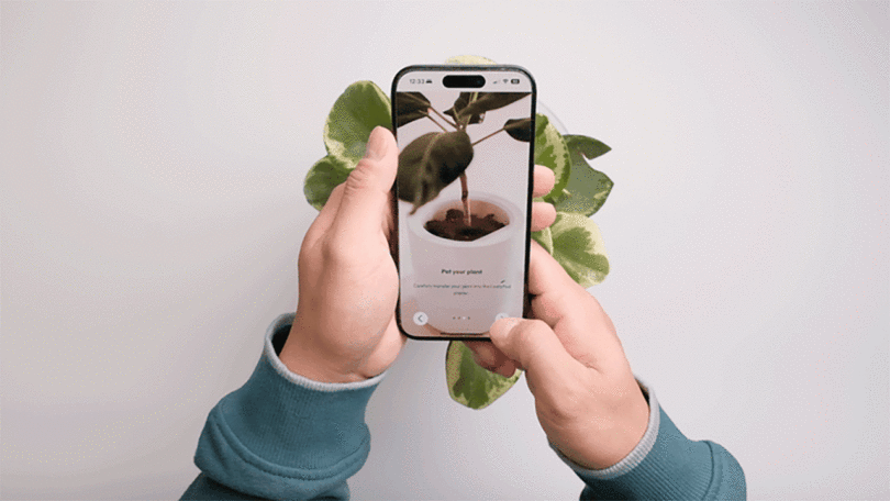 A person holds a smartphone displaying a plant care app, with a real potted plant partially visible behind the phone.