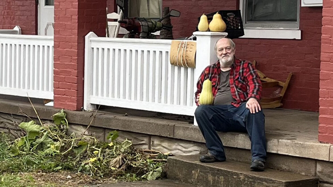 A man in a red plaid shirt sits on porch steps of his housing, holding a squash. Beside him, a sign reads "Free Squash" near a few gourds.