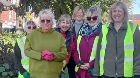 Beverley Urban Gardeners A group of six women wearing fleeces, jumpers, gloves and yellow high vis jackets. They are smiling into the camera and standing near trees and bushes in an urban area.