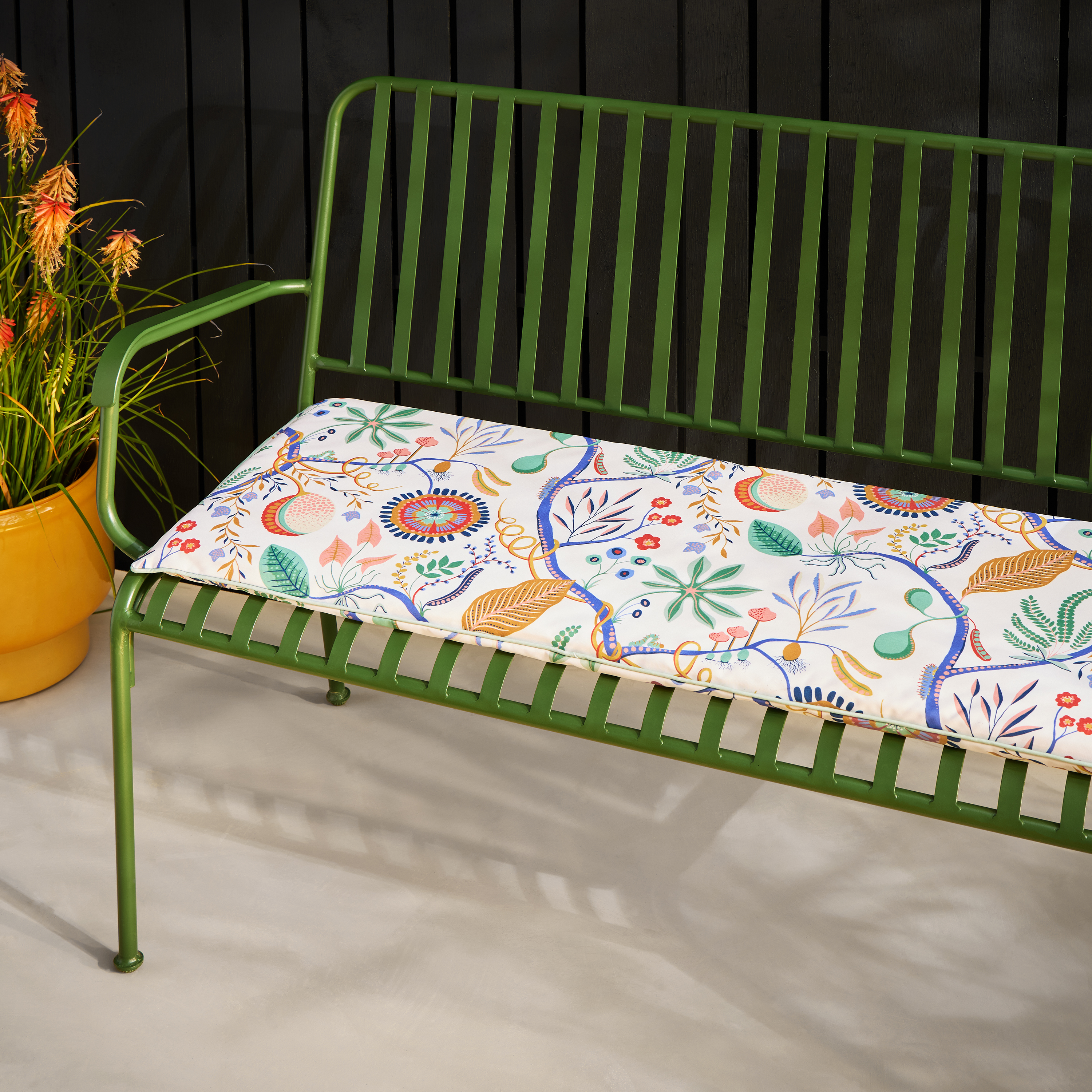 Green metal slatted outdoor bench with a white cushion featuring a colorful floral and botanical pattern.