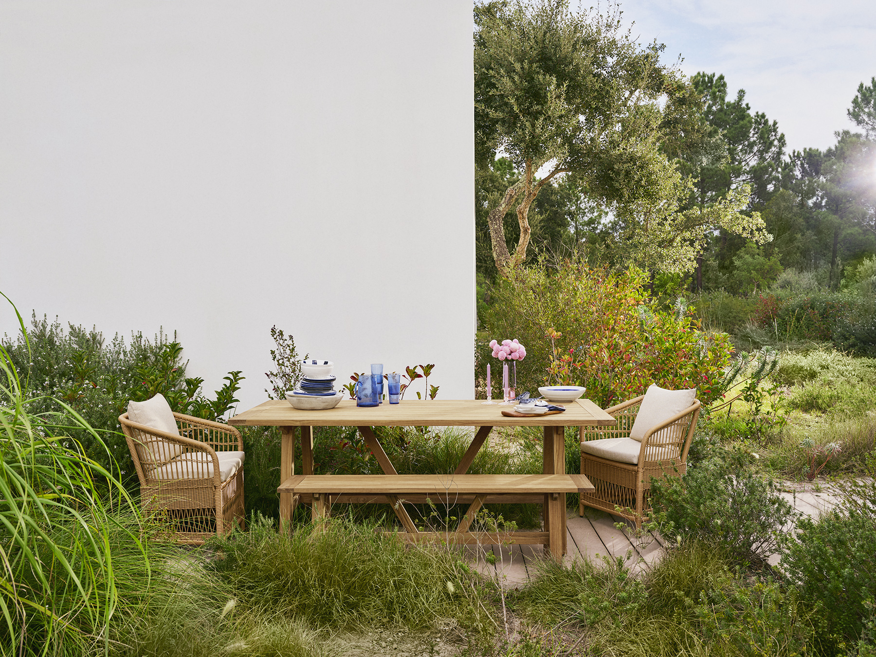 Outdoor dining set with a farmhouse table, bench, and two wicker chairs with cushions, set in a garden with lush greenery and a large tree in the background.