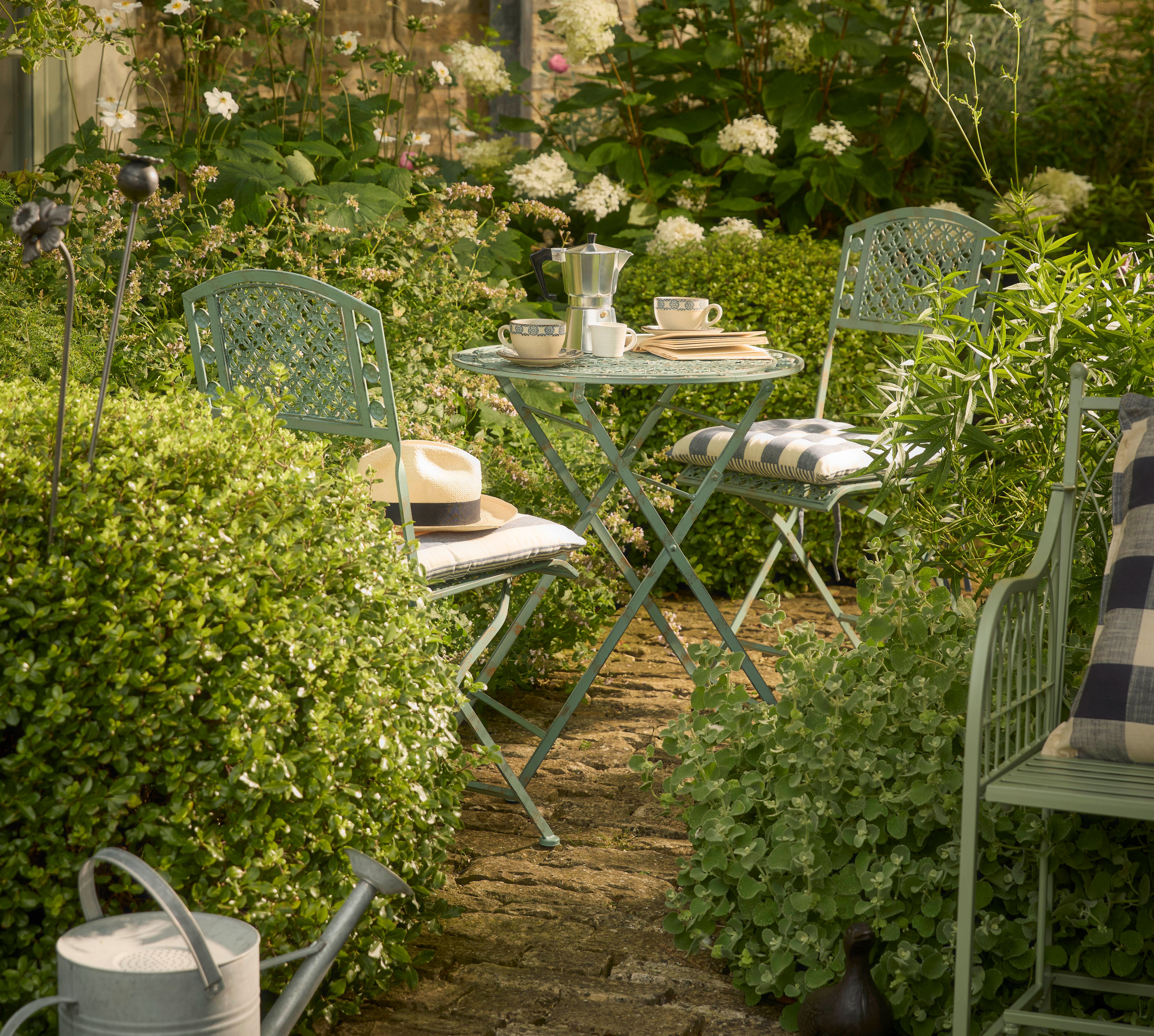 Outdoor bistro set with light green table and two chairs surrounded by lush greenery and flowers.