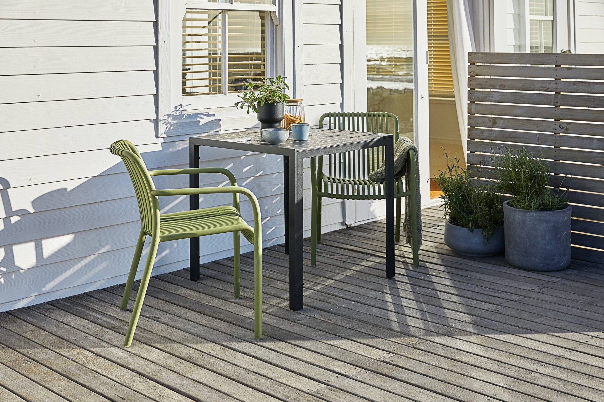 Two olive green JYSK Nabben stacking garden chairs and a small dark wood-look table on a wooden deck next to a white house.