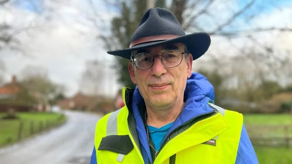 David is wearing a hat has rounded rimmed glasses and a high vise jacket. 