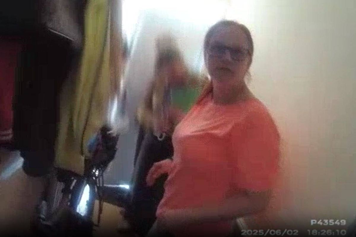 An individual dressed in an orange top stands in a room with a blurred background, where other people are partially visible. The scene is set against a white wall, and the lighting suggests an indoor environment.