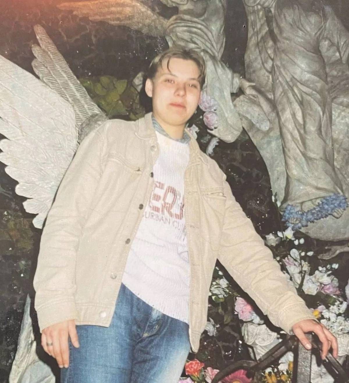 An individual dressed in a beige jacket and a white t-shirt stands in front of a backdrop featuring a large angel statue with expansive wings and intricate detailing. The setting suggests a contemplative or artistic environment.