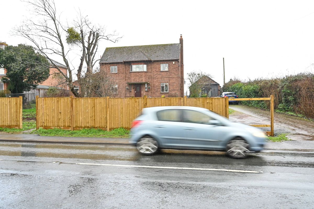 A small blue car is driving down a wet street in front of a wooden fence, with a house visible in the background.