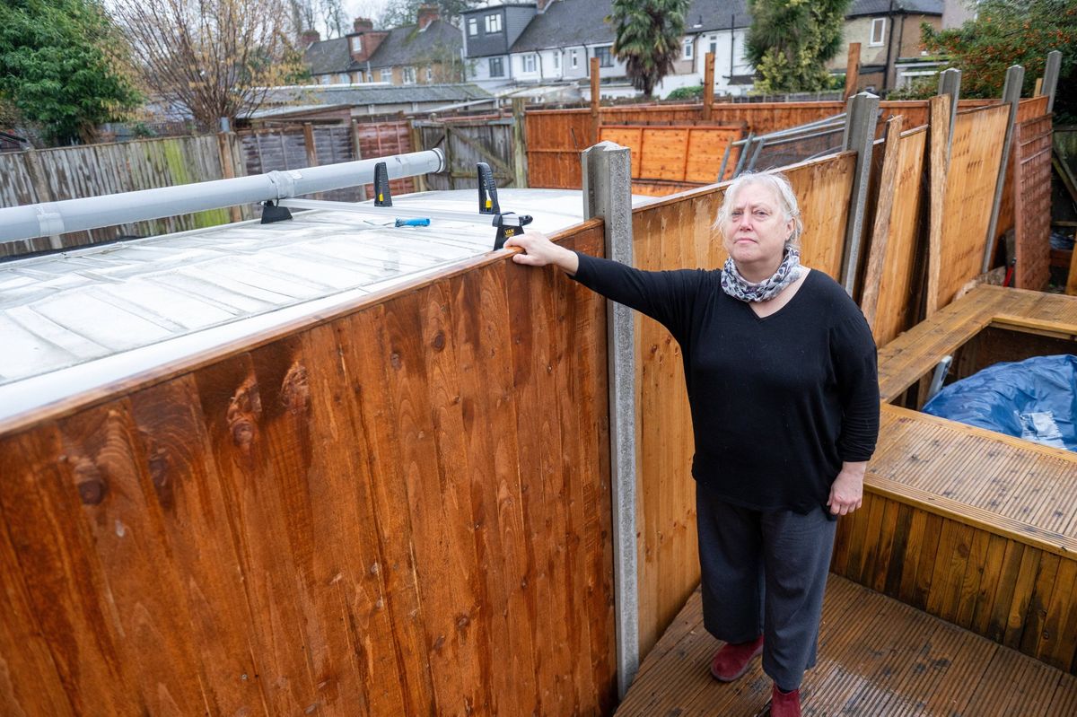 An individual is standing next to a wooden fence, with one hand resting on the top of the fence. The fence appears to be in a state of disrepair, with visible damage and wear. The setting includes a backyard with a house and some greenery in the background. The person is dressed in a dark top and pants, with red boots.