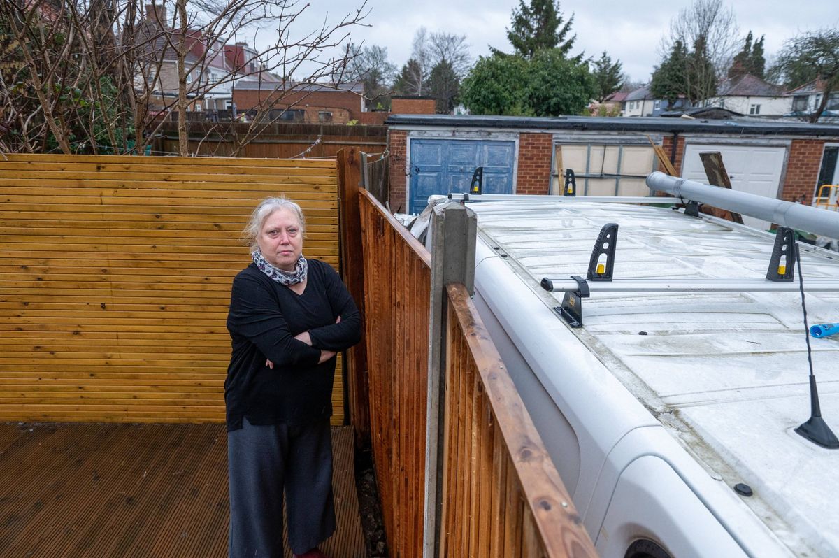 An individual with light hair is standing in a backyard, facing a wooden fence, with a white structure equipped with solar panels in the background.