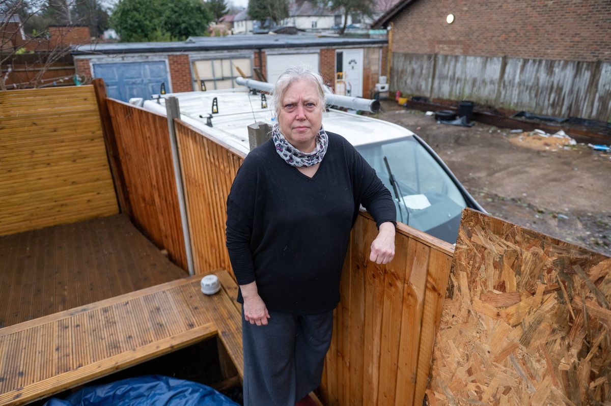 An individual stands in front of a wooden structure, likely engaged in construction or maintenance activities, with a vehicle partially visible in the background.