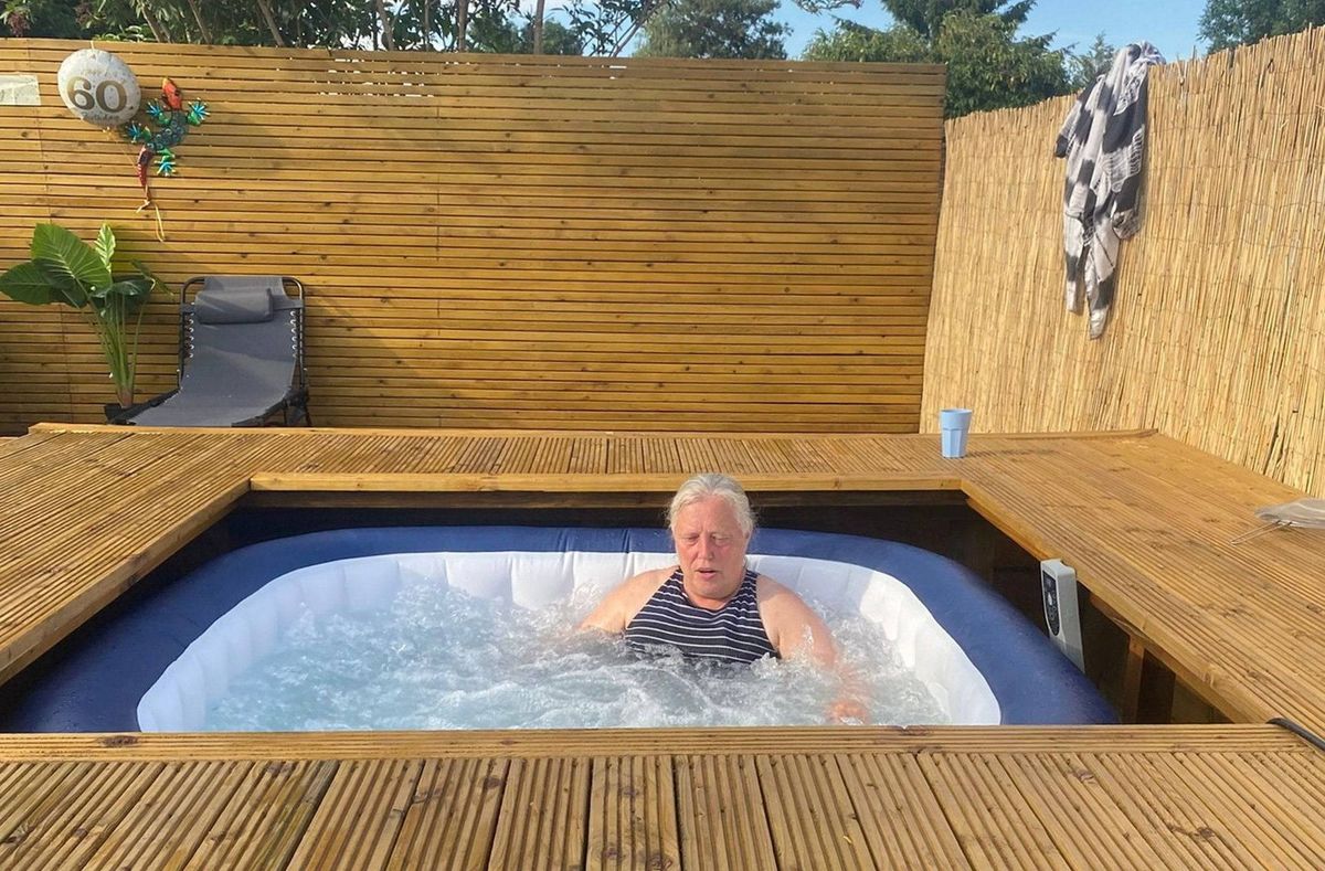An individual is relaxing in an outdoor hot tub, which is enclosed by a wooden structure, with greenery and a towel in the background.