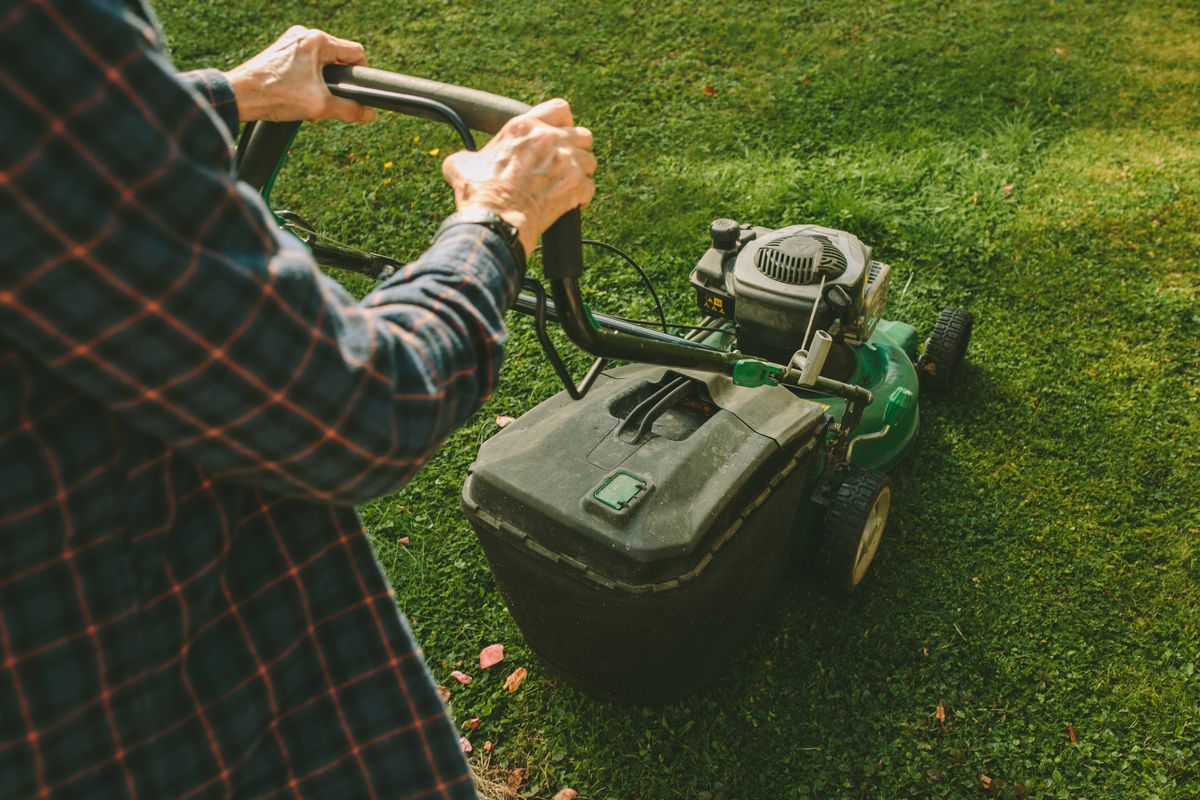 A senior woman operates a push lawn mower in a rustic backyard, surrounded by a mix of green grass, landscaped features, and natural elements. Wearing wellington boots and a plaid shirt, she exemplifies active living and a hands-on approach to gardening and home maintenance. Perfect for themes of gardening, senior lifestyle, and rural living.