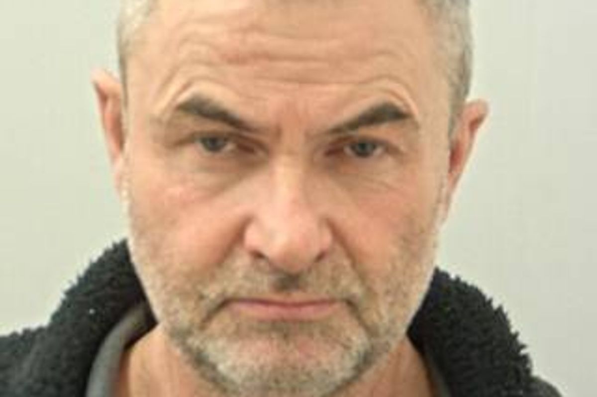 A man with a stern expression, sporting a full beard and mustache, wearing a black jacket, captured in a close-up portrait against a plain background.
