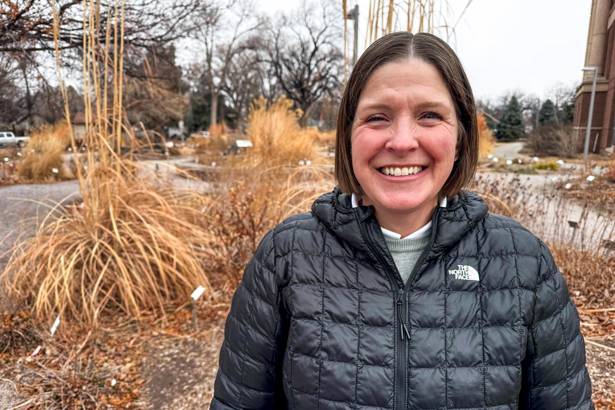 A woman in a black coat smiles at the camera, standing in front of yellow shrubs.