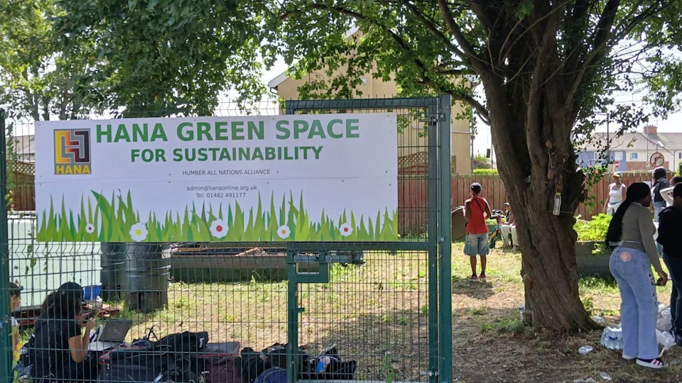 A green area with a green gate in front of it with a poster which says 'HANA Green Space for sustainability' with grass and daisy drawing at the bottom. Various people can be seen inside the space with houses seen in the background.