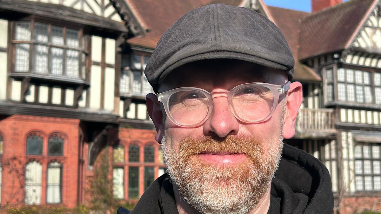 A man with a grey beard and white glasses and a black flat cap is looking at the camera with a slight smile and historic house in the background
